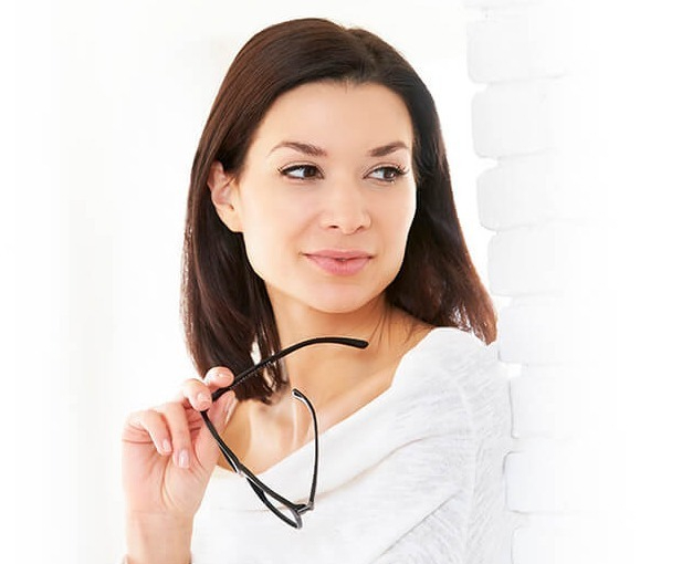 Woman holding eye glasses leaning on wall | Phoenician Eye Specialists